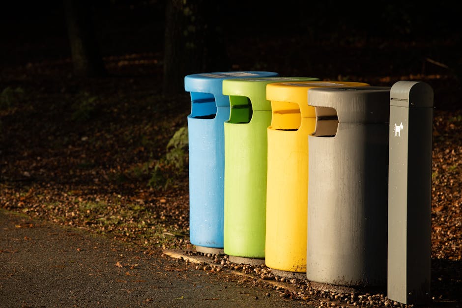 Four cylindrical rubbish bins are aligned on a paved pathway next to a gravel surface in an outdoor setting, each featuring distinct colours and materials. The first bin from the left is light blue with a slightly textured surface, positioned closest to the camera. Next is a bright green bin with a smooth finish, followed by a yellow bin with a similar smooth exterior, and a grey bin with a matte surface. All bins have large, rectangular openings for waste disposal, with the blue and green bins' openings visible at the top, and the yellow and grey bins' openings also facing the viewer. To the right of these bins, a narrow, dark grey waste disposal unit with a small, white dog icon is visible, suggesting it is designated for specific waste types or for use by pet owners. The background features a dark, wooded area with minimal natural light, highlighting the bins against the subdued environment, which is typical for private or independent waste collection points associated with rubbish removal services researching alternative waste handling options in urban outdoor spaces.